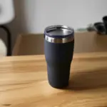 A sleek, modern stainless steel travel mug with a leak-proof lid, resting on a wooden table.