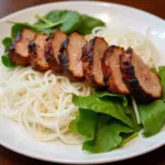 A close-up shot of a plate of Bun Cha, showcasing the grilled pork, vermicelli noodles, and fresh herbs.